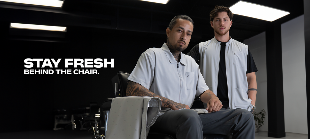 Stay fresh behind the chair. Two barbers in the Cool Gray Collection: one seated in a barber chair in The Barber Jacket and Barber Pants, the other standing in The Barber Vest, with a gray Barber Cape draped over the chair arm.